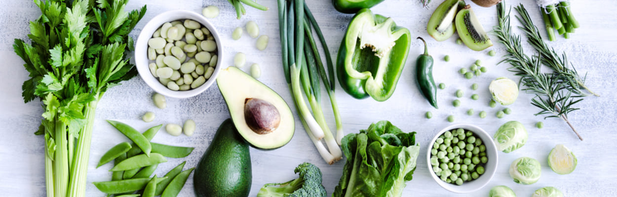 Em uma superfície clara, vários alimentos verdes estão dispostos espalhados, como abacate, brócolis, ervilha, kiwi, couve.