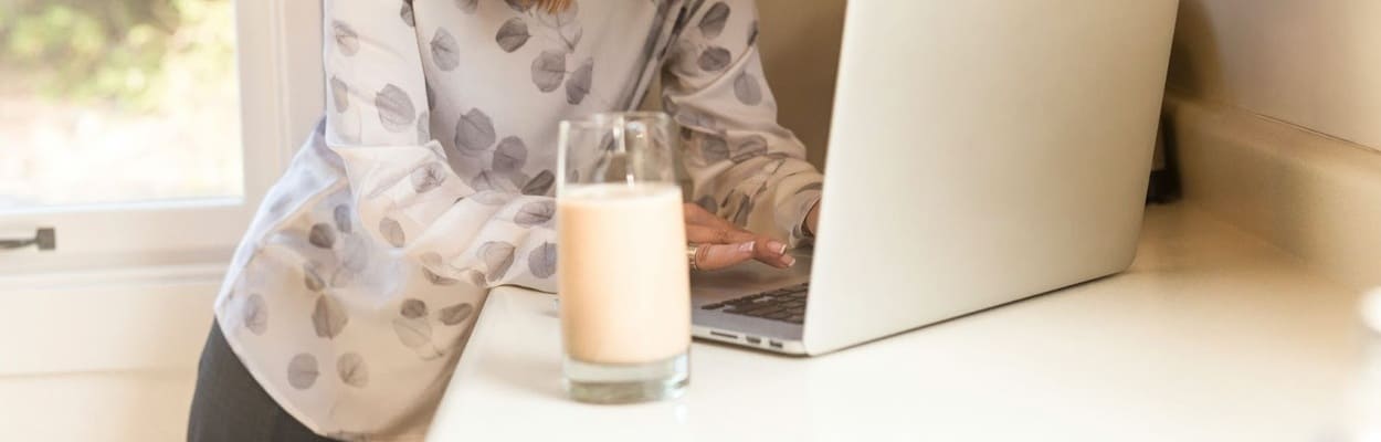 Mulher em pé em um balcão de cozinha com um notebook aberto e um copo com leite pesquisando as proteínas do leite.