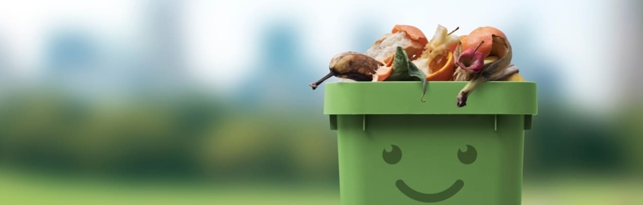 Caixa de lixo verde, com um desenho de sorriso, cheia de resíduos orgânicos biodegradáveis.