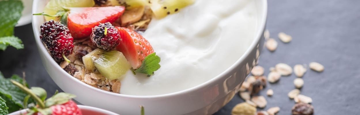 Close-up de uma tigela posicionada em uma mesa, repleta dos benefícios do iogurte natural acompanhado de granola e uma variedade de frutas por cima.