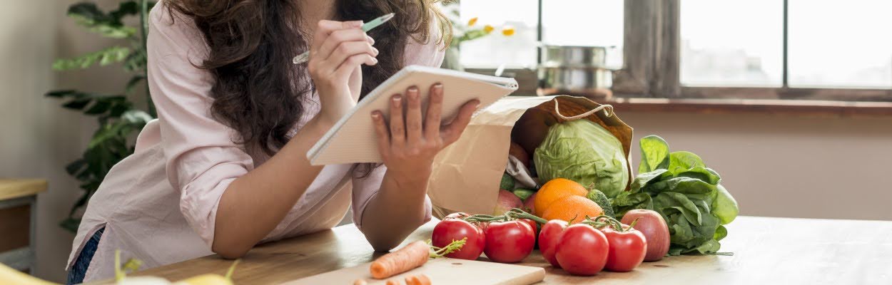 Mulher branca de cabelos castanhos está segurando um caderno, apoiada na bancada da cozinha rodeada por legumes e verduras, anotando dicas de trocas saudáveis. 