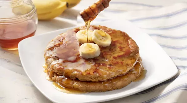 Em um prato branco, há duas panquecas de banana e pasta de amendoim e um pote de mel ao lado.