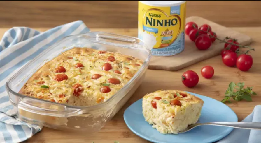 Torta de frango cremosa com Ninho em uma travessa transparente. Ao redor, vemos um prato raso azul com uma porção da torta, tomates-cereja e uma lata de leite Ninho.
