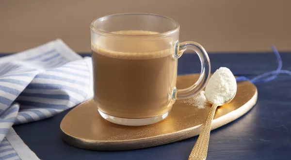 Fotografia em tons de marrom em uma bancada de madeira de cor azul. Ao centro, uma tábua de madeira dourada contendo uma xícara de café com uma colher de leite em pó ao lado. Ao fundo, há um pano listrado azul.