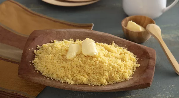 Foto da receita de Cuscuz de Milho. Observa-se uma tigela de madeira com o cuscuz soltinho e com manteiga em cima derretendo.