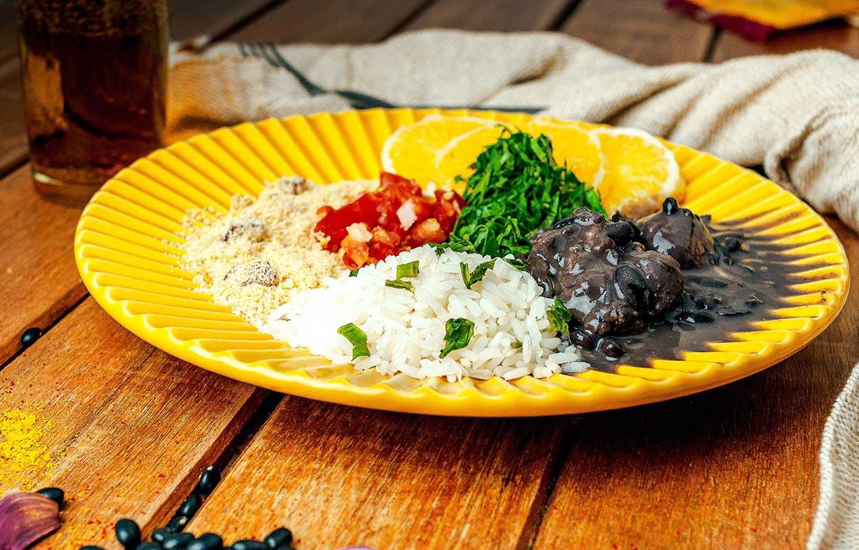 prato amarelo em cima de uma mesa de madeira com arroz, feijão preto, farofa, tomate e couve