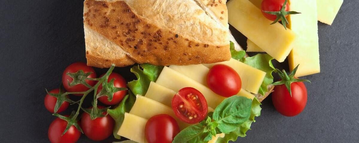 Vista aérea de ingredientes para um lanche nutritivo: pão com gergelim, mini tomates, alface e fatias de queijo mussarela.
