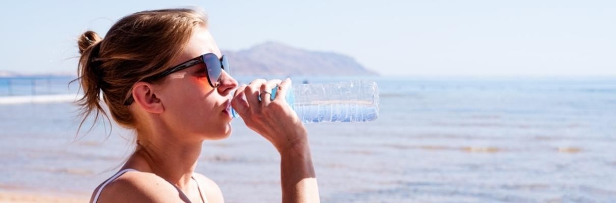Mulher com óculos escuro bebendo uma garrafa de água em frente à uma praia tropical.