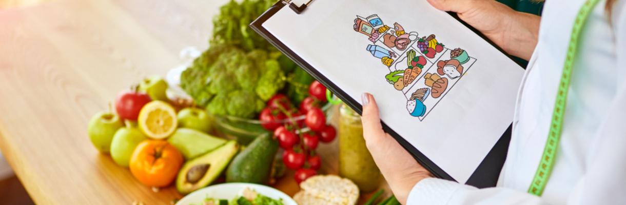 Foto de uma nutricionista segurando uma prancheta que contém a ilustração de uma pirâmide alimentar e em sua frente há uma mesa de madeira com vários alimentos, como frutas, legumes e verduras.