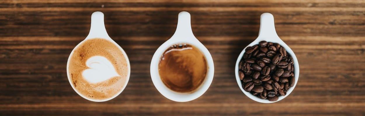 Em uma mesa de madeira, há três pequenas xícaras de café. Da esquerda para direita, temos uma com o desenho de coração feito com leite, o do meio é um expresso, e o da direita possui o café em grão.