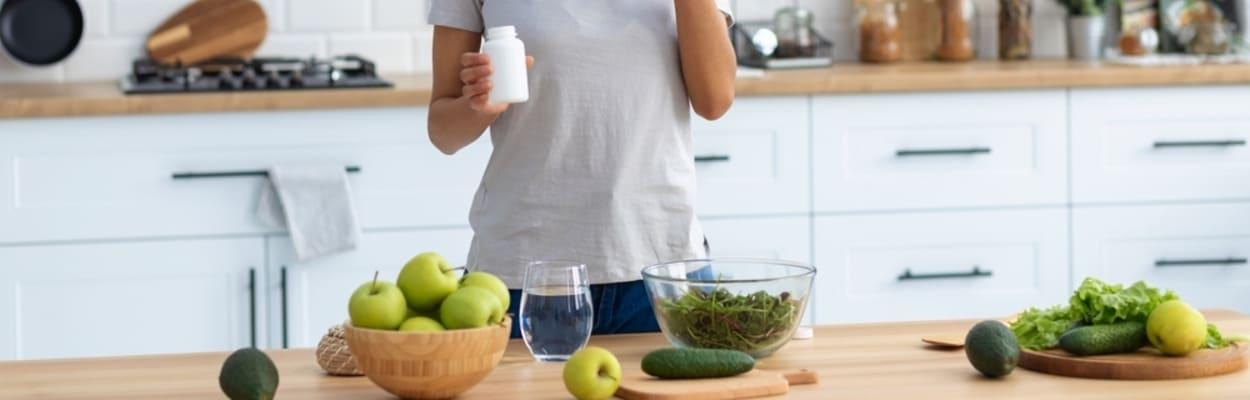 Em uma cozinha, uma mulher negra, de cabelos crespos presos em um coque, toma suas vitaminas para o corpo feminino e em sua frente há uma bancada com frutas e verduras verdes.