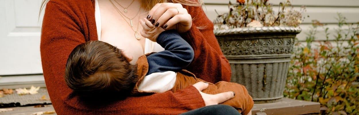 Uma mulher branca, de cabelos longos loiros, está amamentando seu bebê, representando a alimentação na amamentação.