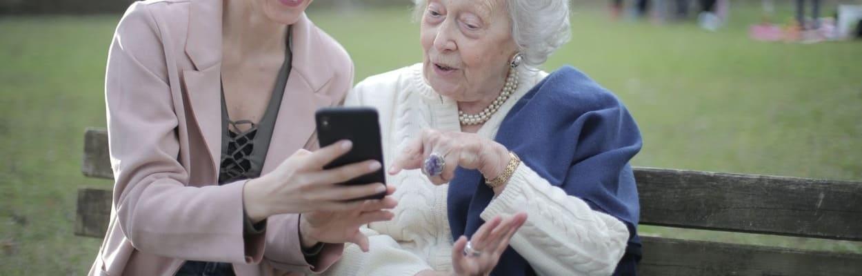 Uma mulher jovem e uma idosa saudáveis, sentadas em um parque olhando um celular.