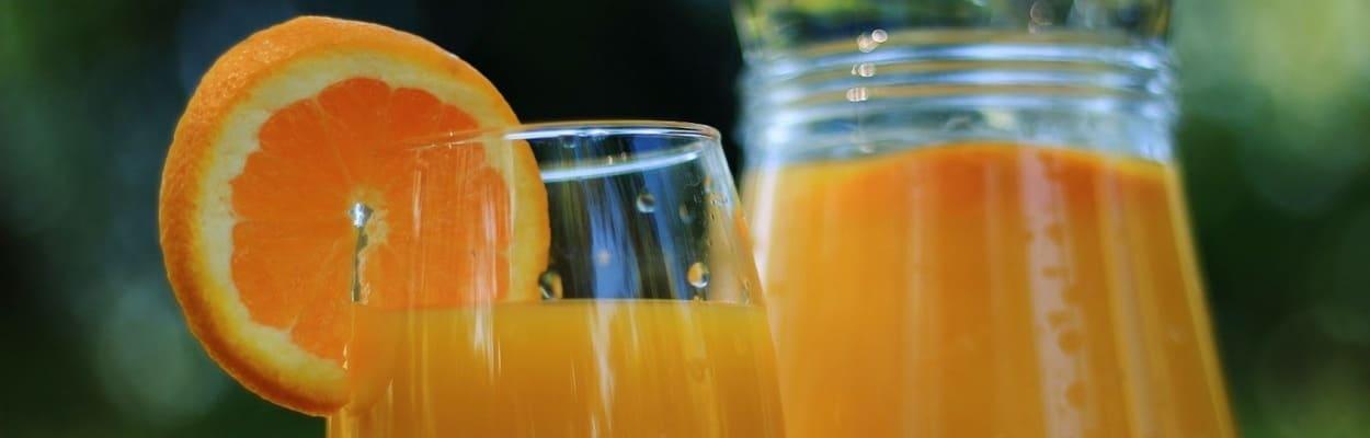 Copo de vidro com suco de laranja, com uma fatia de laranja na borda e uma jarra de suco de laranja atrás.