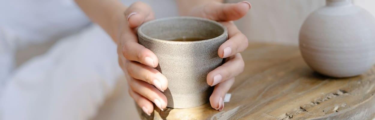 Pessoa com braços esticados pegando um copo cinza com chá em uma mesinha com vaso em cima.