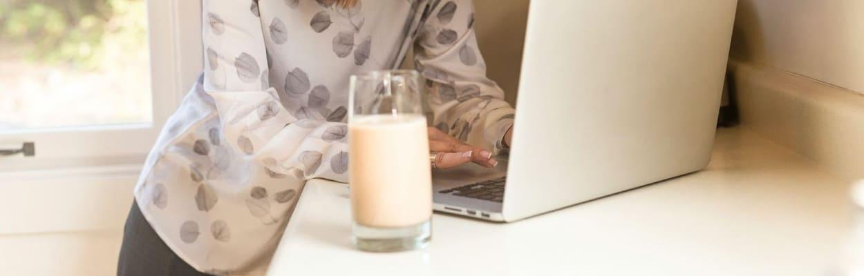 Mulher em pé em um balcão de cozinha com um notebook aberto e um copo com leite pesquisando as proteínas do leite.