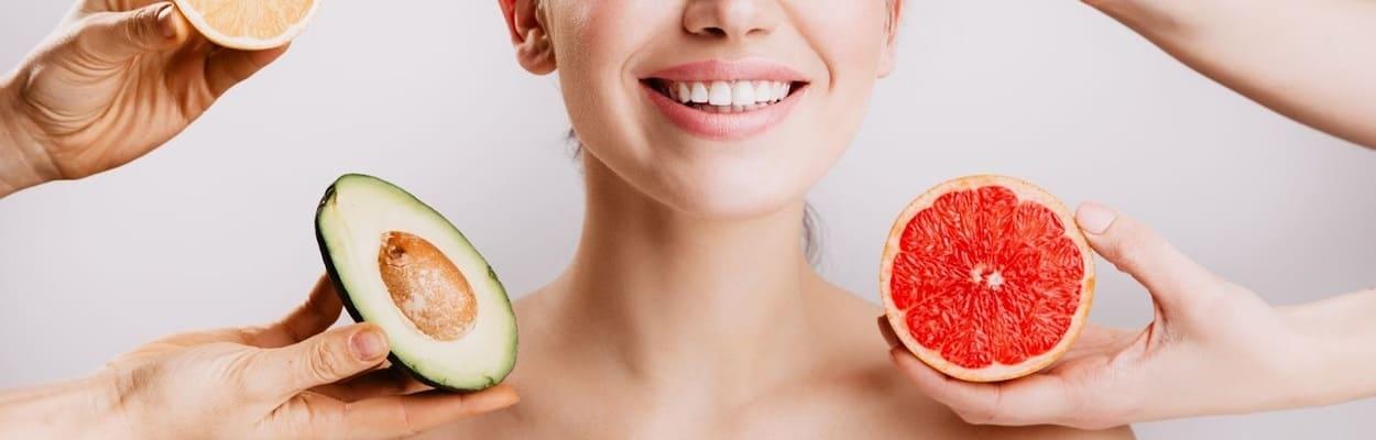 Mulher sorrindo num fundo branco, com mãos de outras pessoas ao seu redor com frutas ricas em vitaminas para pele.