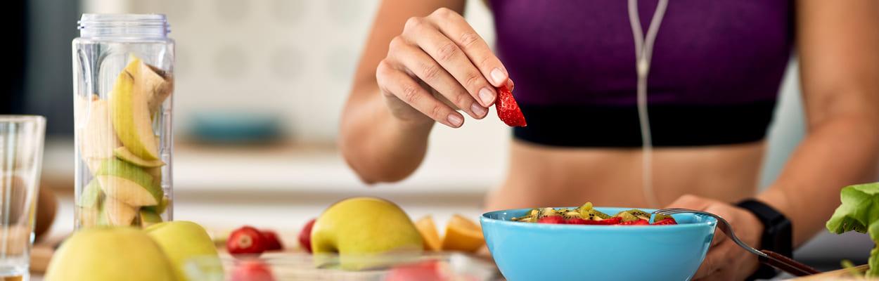 Mulher com roupa de academia adicionando morangos em um pote de salada de frutas. A imagem representa o que comer antes do treino para ganhar massa muscular.