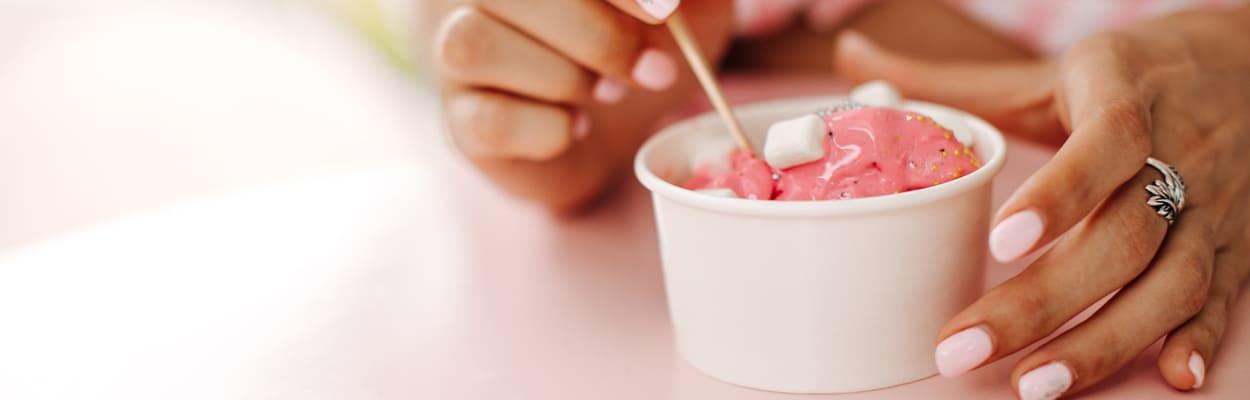 Mãos de uma mulher segurando um pote branco de sorvete com a cor rosa. A imagem representa a dúvida se tomar sorvete gripado faz mal.  