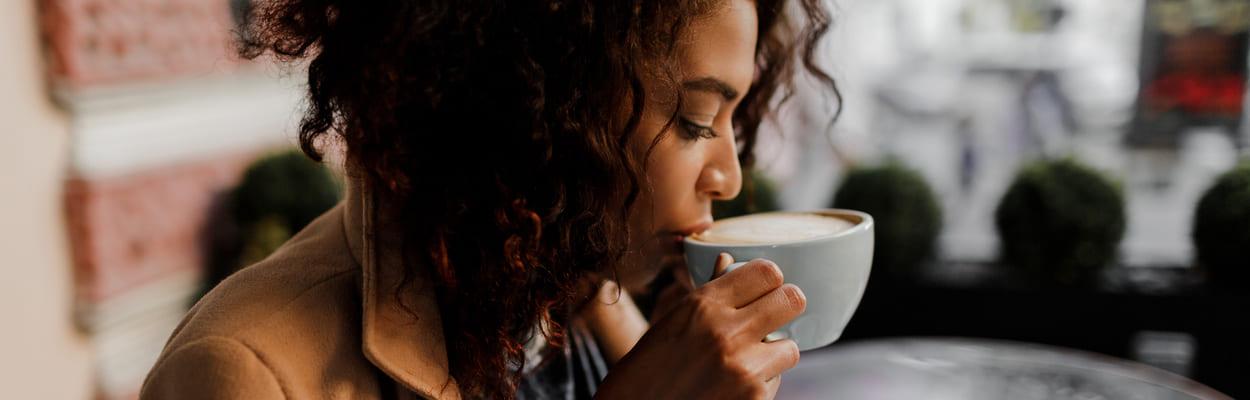 Mulher com blusa de frio sentada enquanto toma uma xícara de café. A imagem representa o que é café descafeinado.