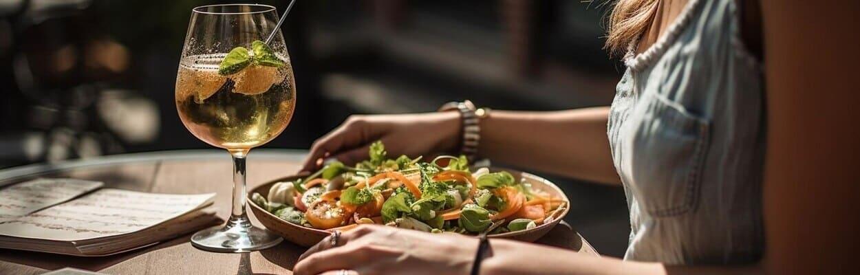 Mulher com as mãos apoiadas na mesa. Em sua frente, vemos um prato repleto de salada e uma taça com bebida. A imagem representa o jantar para os dias quentes.