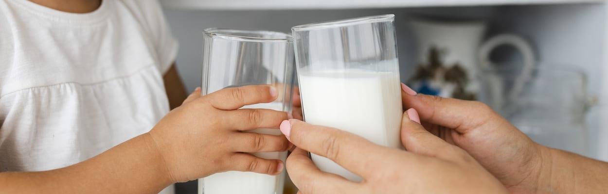 Mãos de uma mulher adulta e de uma criança, cada uma segura um copo de leite. A imagem representa os sintomas da intolerância à lactose.