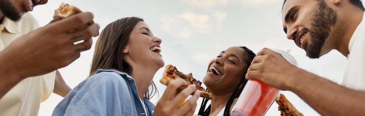 Jovens dando risada enquanto comem um pedaço de pizza e seguram copos. Eles estão se divertindo em um festival. A imagem representa o que levar para comer em um festival.