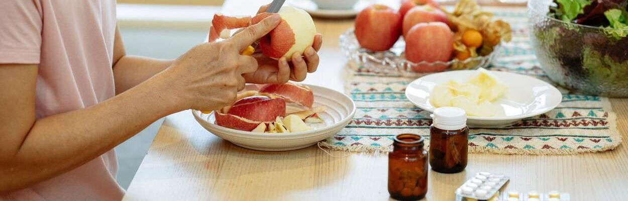 Pessoa passando por um pós-cirúrgico descascando uma maçã, numa mesa com outras frutas e remédios.