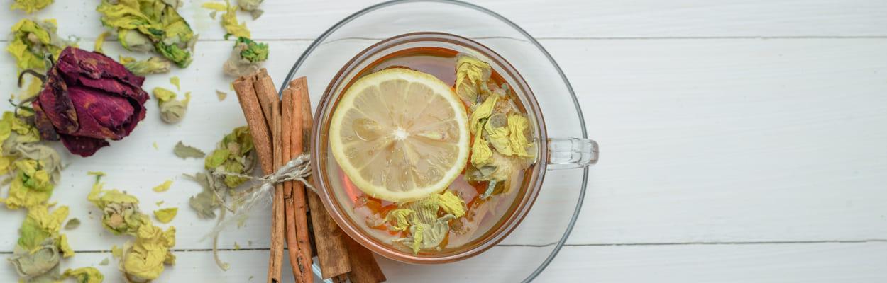 Xícara de chá transparente com infusão de ervas, uma rodela de limão de canela em pau. A imagem representa os chás para dieta alcalina.