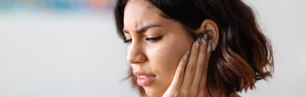 Mulher de cabelo curto e camiseta laranja está com a mão no ouvido, enquanto faz expressão de dor. A imagem representa o que é bom para a dor de ouvido.
