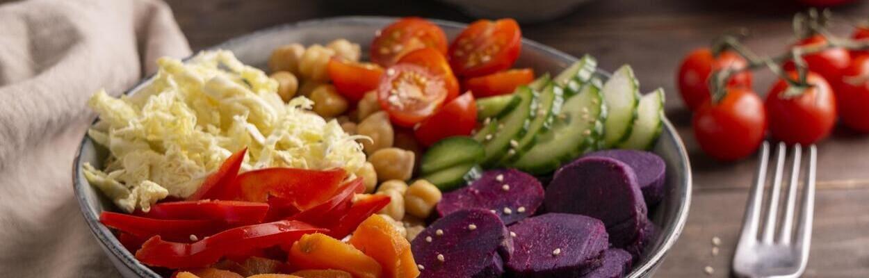 Tigela cinza com legumes dentro, dentre eles estão cenouras fatiadas, tomates, pepinos e grão-de-bico. A imagem representa uma dieta à base de vegetais.