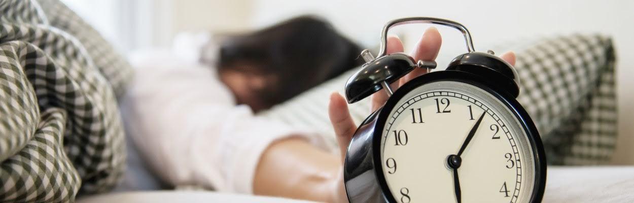 Deitada em uma cama, uma mulher está desligando um despertador antigo, preto e branco, que vemos em primeiro plano, ilustrando qual a melhor hora para dormir e acordar.